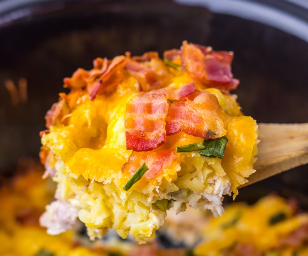 Close-up of a scoop of cheesy casserole with bacon and green onions, held on a wooden spoon.