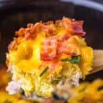 Close-up of a scoop of cheesy casserole with bacon and green onions, held on a wooden spoon.