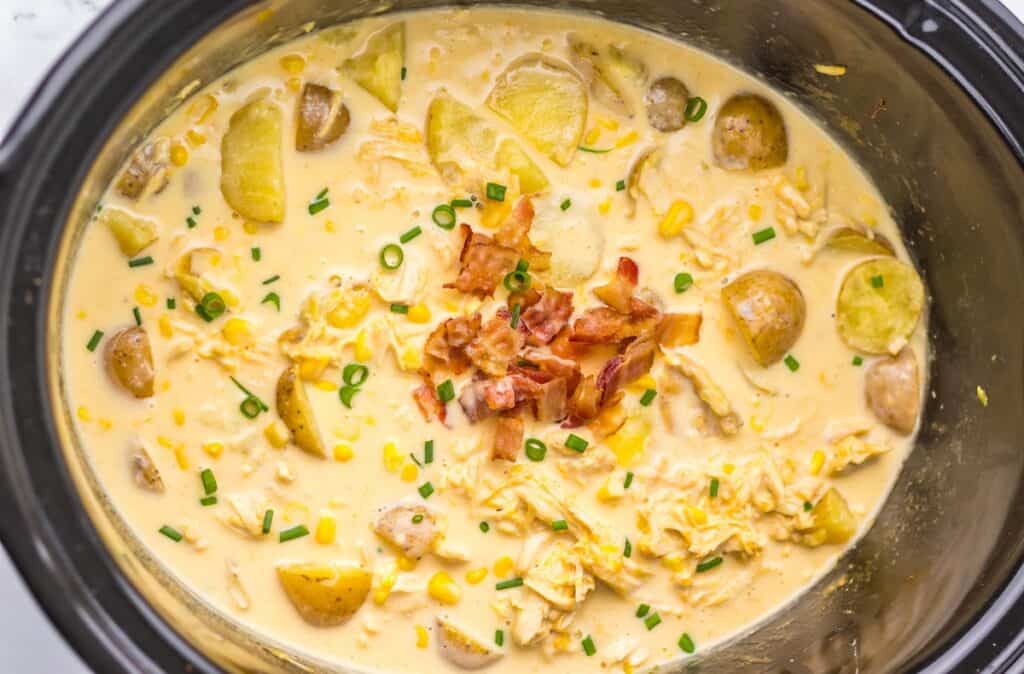 Creamy soup with chicken, potatoes, corn, and cheese in a slow cooker, topped with chopped bacon and green onions.