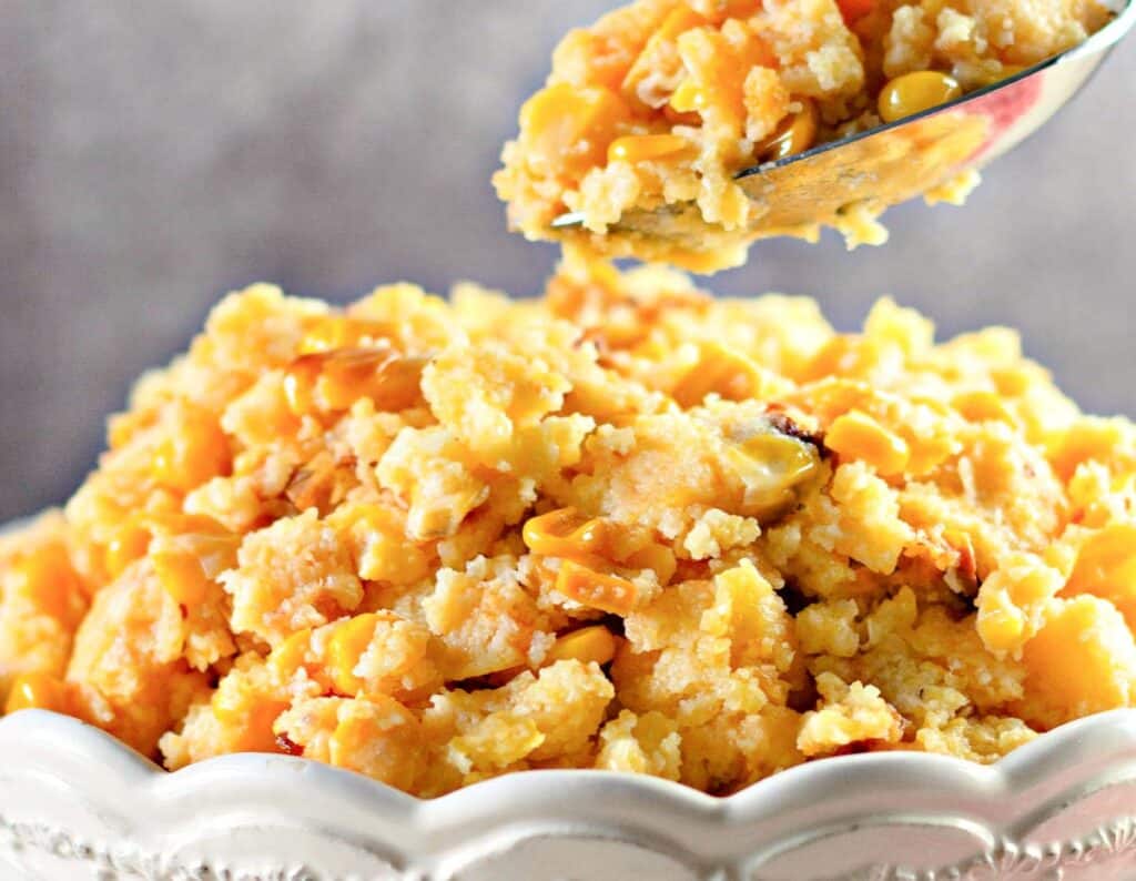 A close-up of a bowl of corn casserole with a serving spoon lifting a portion. The casserole has a golden, crumbly texture.