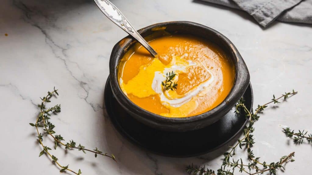 A bowl of creamy pumpkin soup with a swirl of cream, garnished with a sprig of thyme. A silver spoon rests in the bowl on a dark plate.