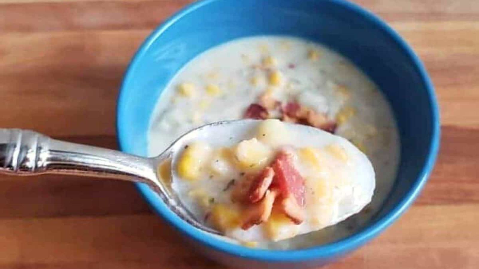 A spoonful of corn chowder with bacon is held over a blue bowl on a wooden surface.