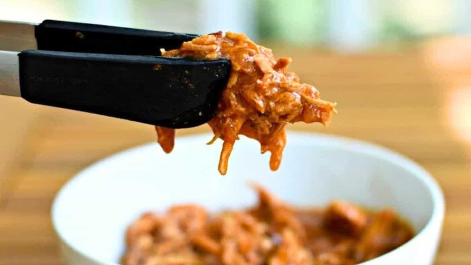 Tongs holding shredded barbeque pork over a white bowl with more pork inside.