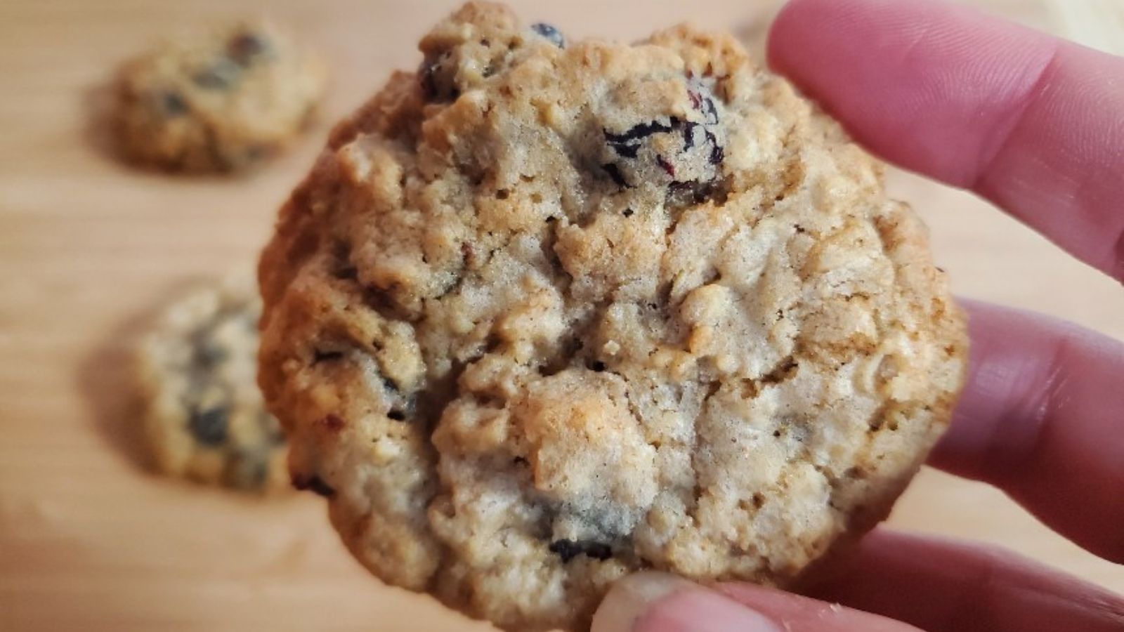 Hand holding an oatmeal raisin cookie with more cookies in the background on a wooden surface.