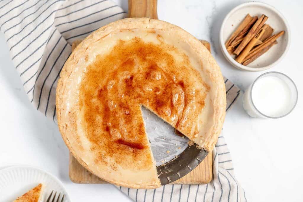 A custard pie with a slice removed on a wooden board, next to a bowl of cinnamon sticks and a glass of milk.