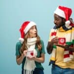 A man and woman wearing Santa hats and scarves hold mugs, exchanging surprised expressions against a blue background.