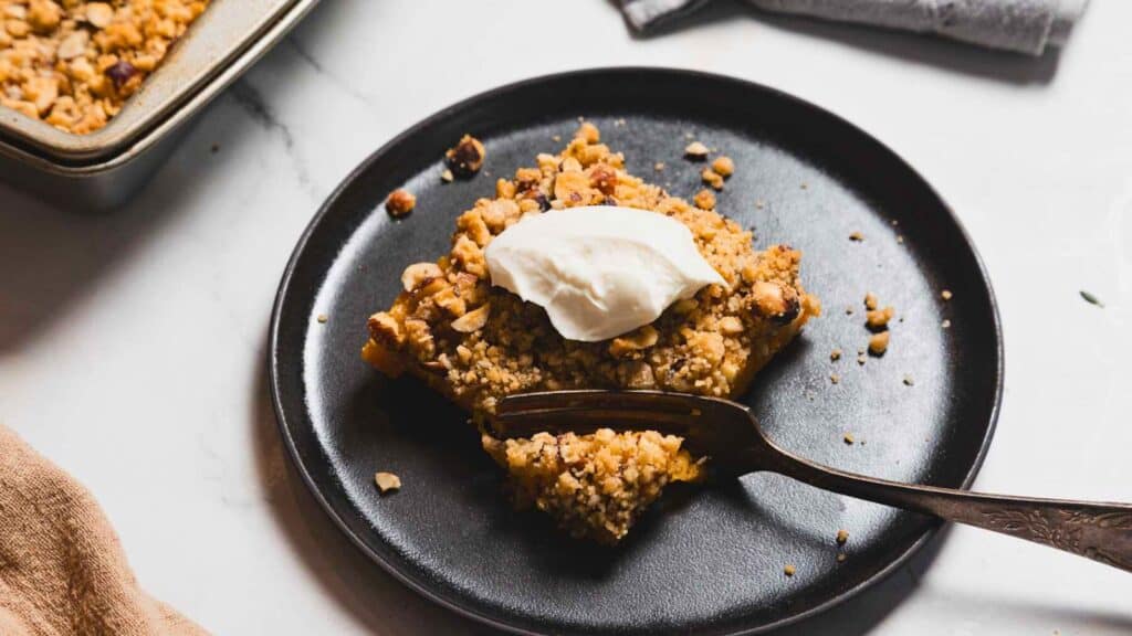 A crumbly dessert square topped with whipped cream on a black plate, with a fork beside it.