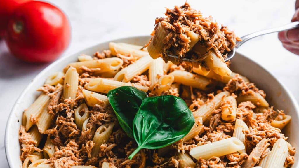 A bowl of pasta with shredded meat and a basil leaf on top. A fork is lifting some pasta and meat from the bowl. Two tomatoes are in the background.