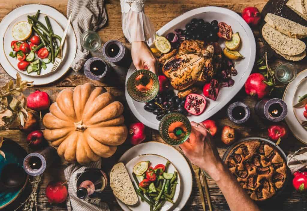 Table with a roasted chicken centerpiece, surrounded by fruits, vegetables, bread, pumpkin, and desserts. People clink glasses over the meal.