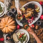 Table with a roasted chicken centerpiece, surrounded by fruits, vegetables, bread, pumpkin, and desserts. People clink glasses over the meal.