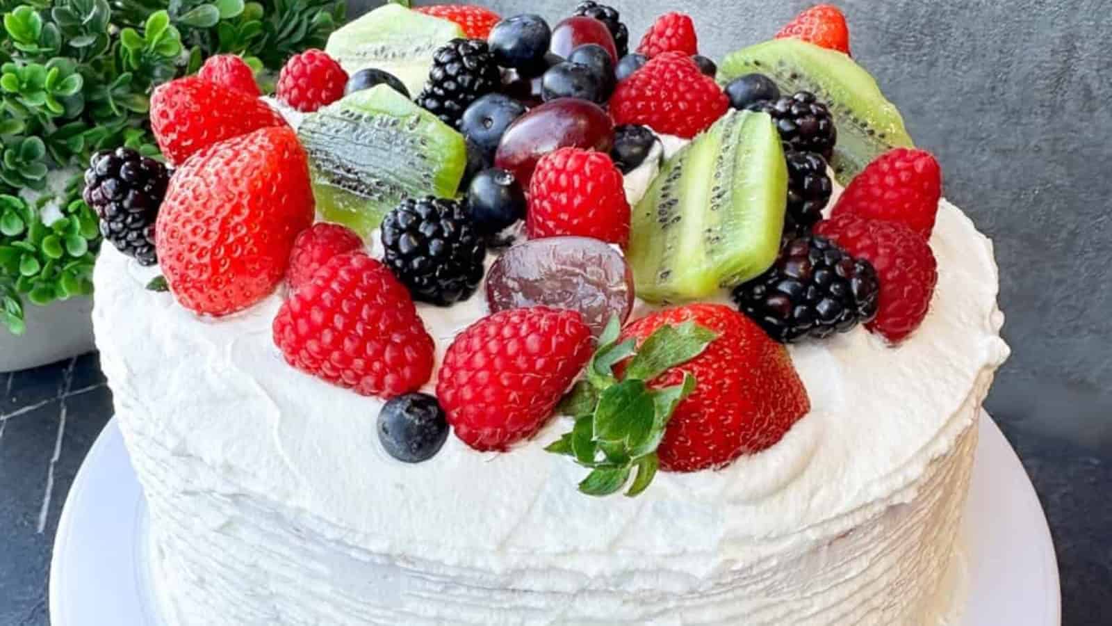 A whipped cream-covered cake topped with strawberries, raspberries, blackberries, blueberries, kiwis, and grapes on a white plate.