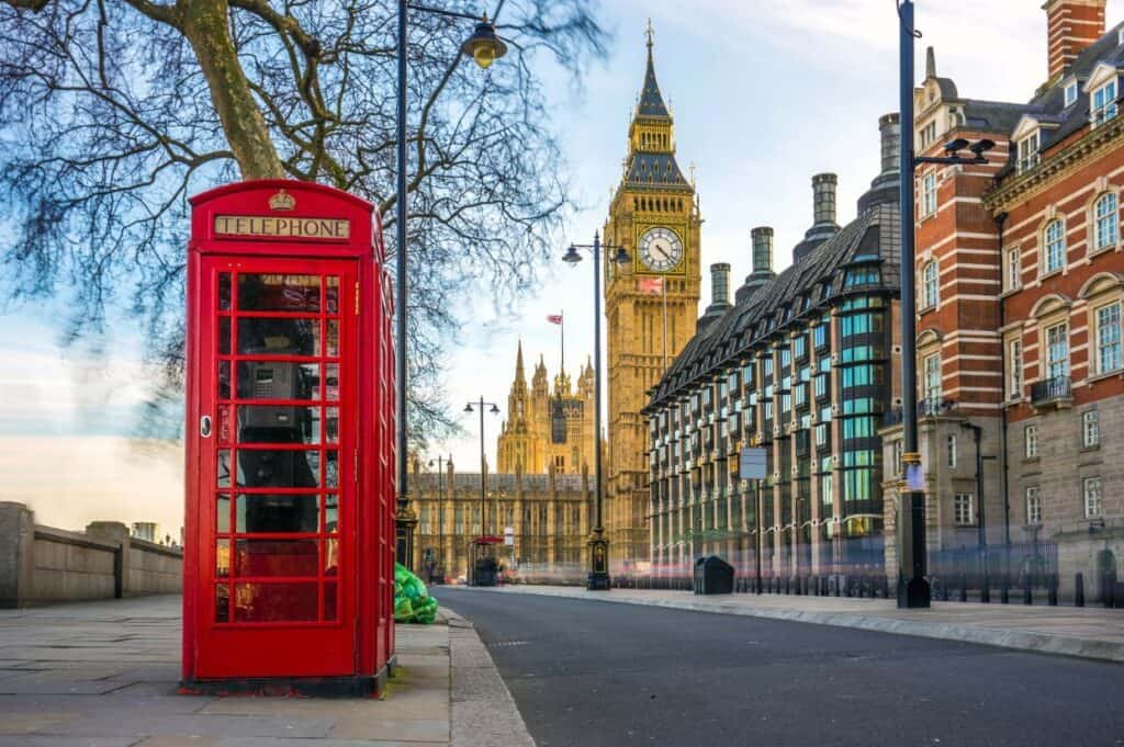 On a bustling street, a classic red telephone booth stands proudly with Big Ben and the Houses of Parliament in the backdrop, capturing the essence of London—a perfect start for memorable day trips from London.