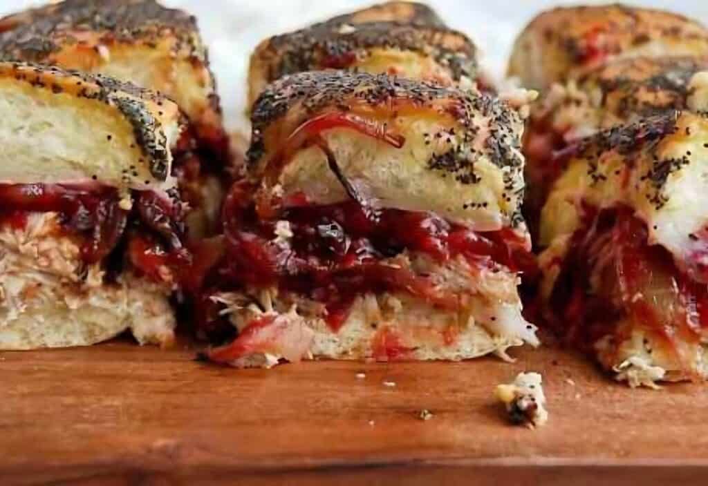 Close-up of sliced turkey sliders with poppy seed crust, filled with cranberry sauce and cheese, displayed on a wooden board.