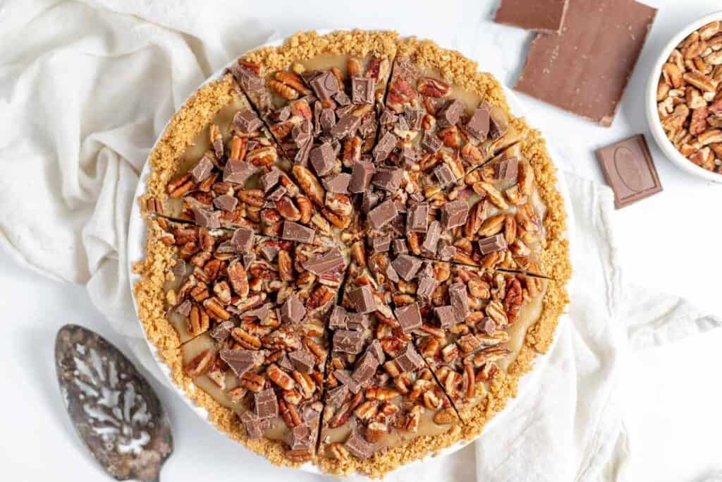 A pecan pie topped with chopped chocolate pieces, sliced into eight portions, on a white cloth with chocolate pieces and pecans nearby.