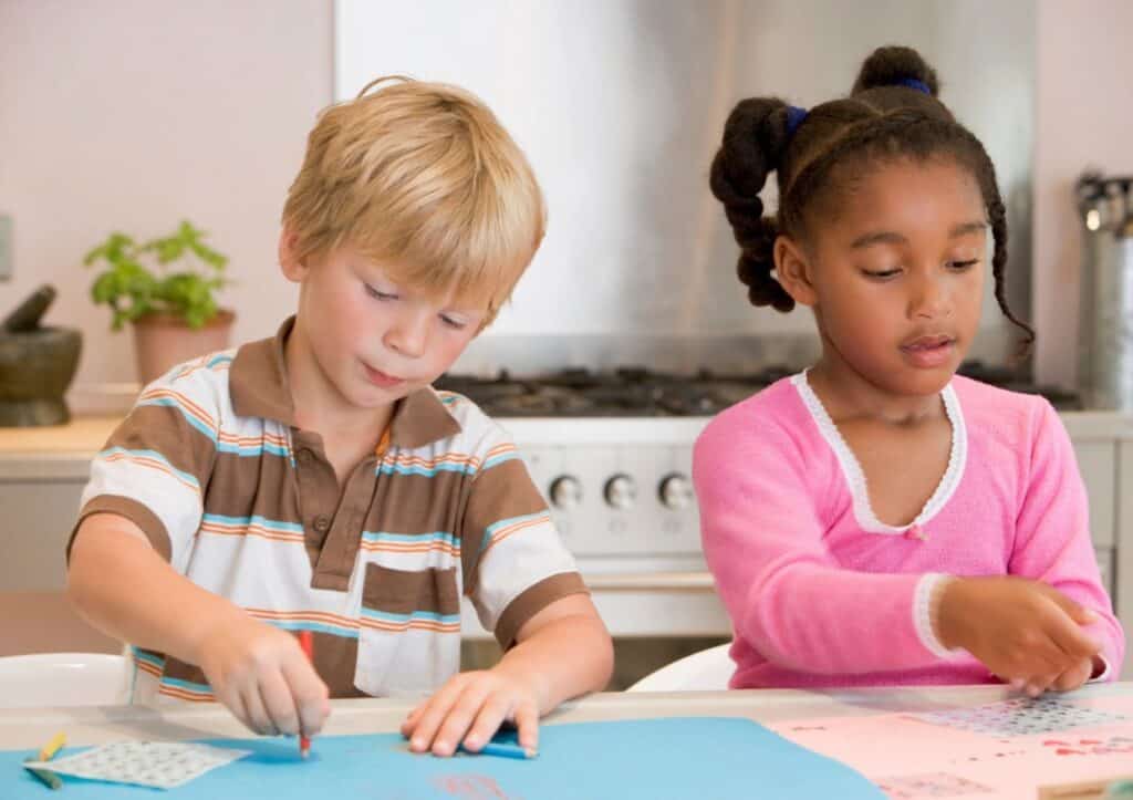 Two children are seated at a table engaged in arts and crafts activities. One is drawing on blue paper, and the other is working on pink paper. They appear focused on their tasks.