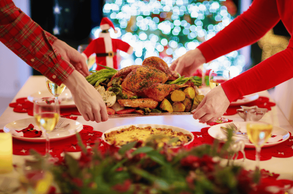 Hands placing a platter of roasted chicken, potatoes, and asparagus on a festive table with a holiday backdrop, inviting you to enjoy the holidays guilt-free.