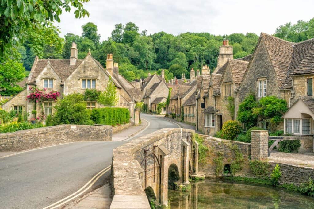 A stone bridge leads to a picturesque village with stone cottages and lush greenery under a cloudy sky, making it an ideal spot for memorable day trips from London.