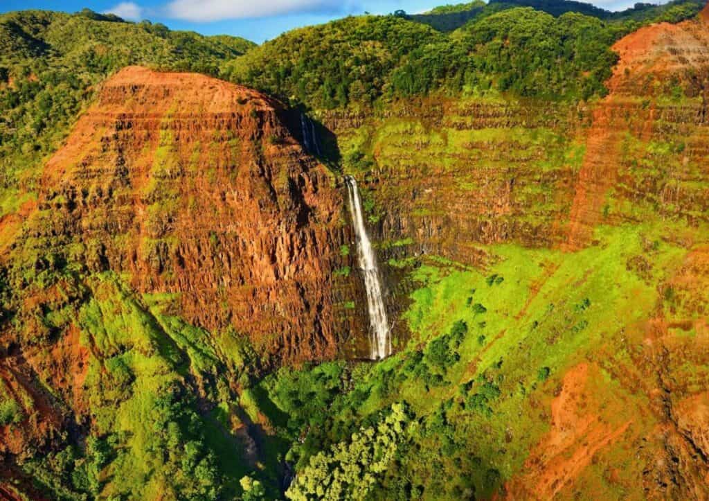 A waterfall cascades down a red and green cliff in a lush, mountainous landscape, offering one of the many breathtaking experiences among the things to do in Kauai.