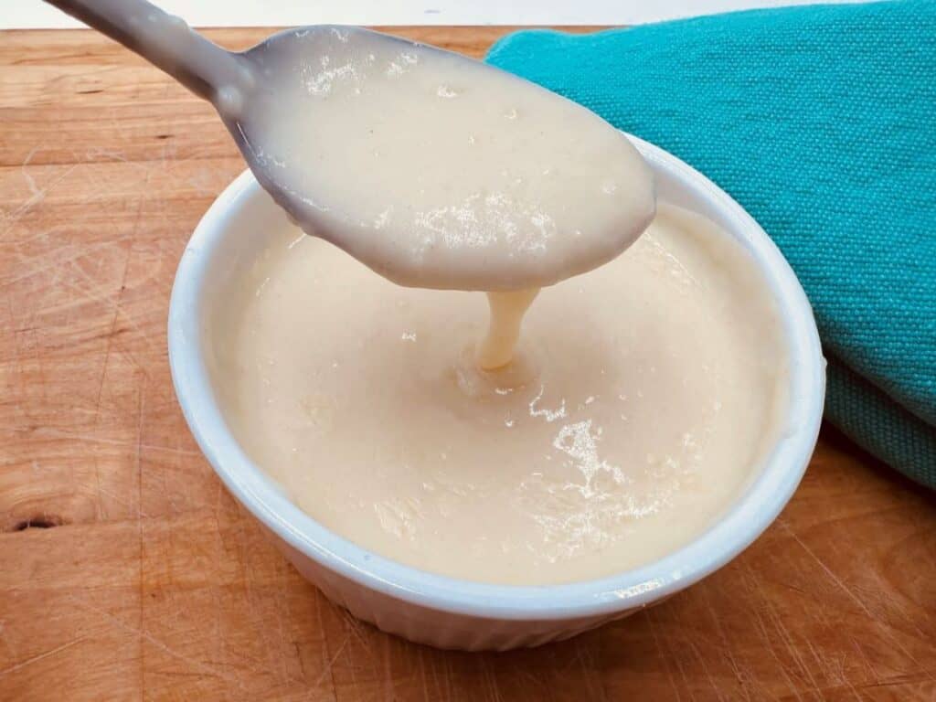 A spoon drips white gravy into a small white ramekin on a wooden surface, with a teal cloth nearby.