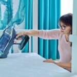 A person demonstrates how to clean a mattress by using a handheld vacuum cleaner, set in a room featuring teal curtains and vibrant cactus wall art.