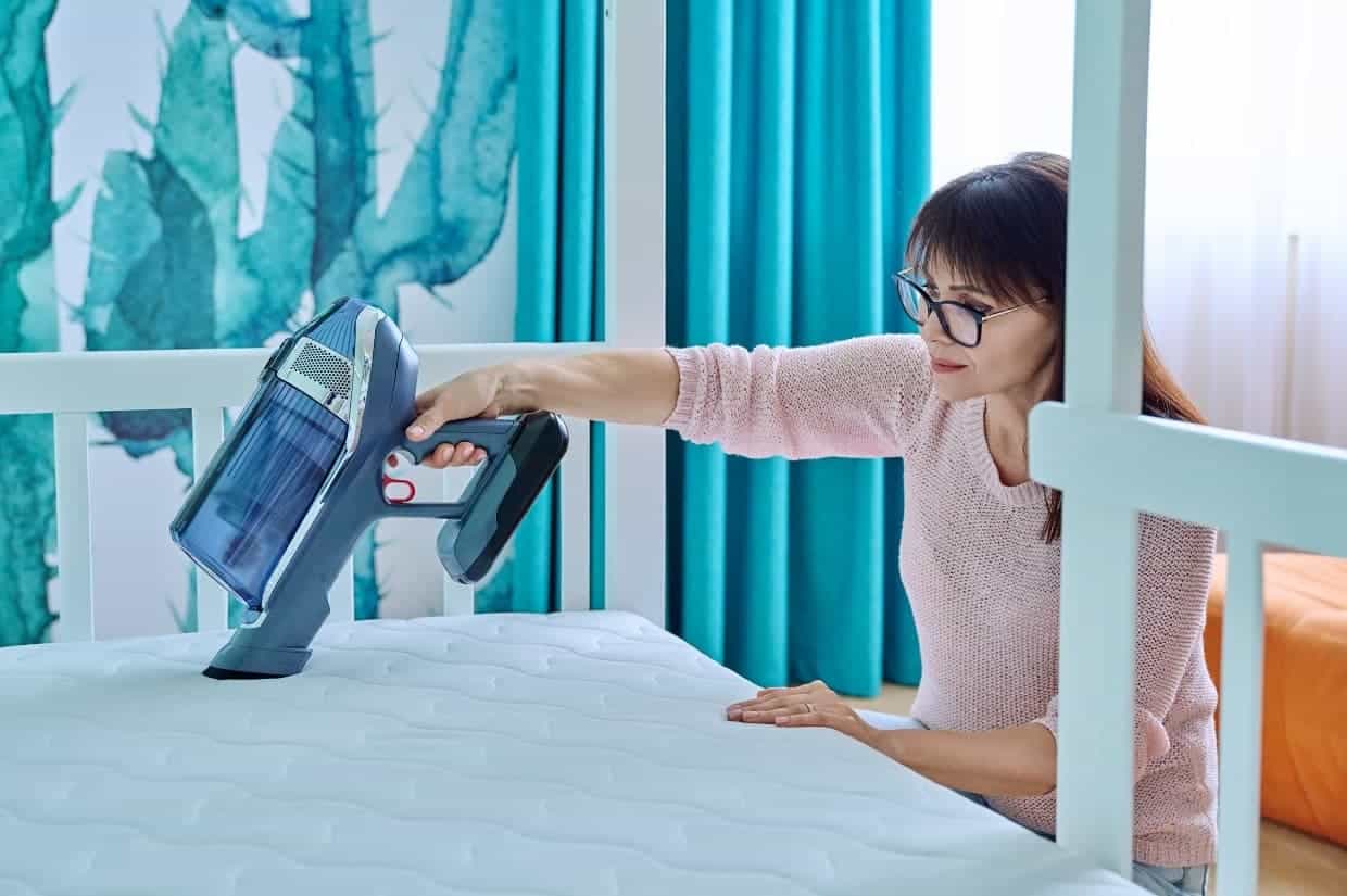 A person demonstrates how to clean a mattress by using a handheld vacuum cleaner, set in a room featuring teal curtains and vibrant cactus wall art.