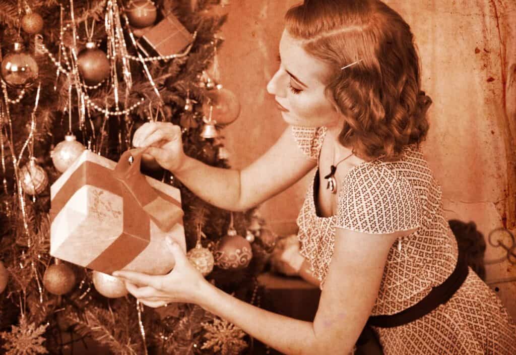 A woman decorates a Christmas tree, placing a gift beneath it. The image has a sepia tone, giving it a vintage look.