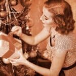 A woman decorates a Christmas tree, placing a gift beneath it. The image has a sepia tone, giving it a vintage look.