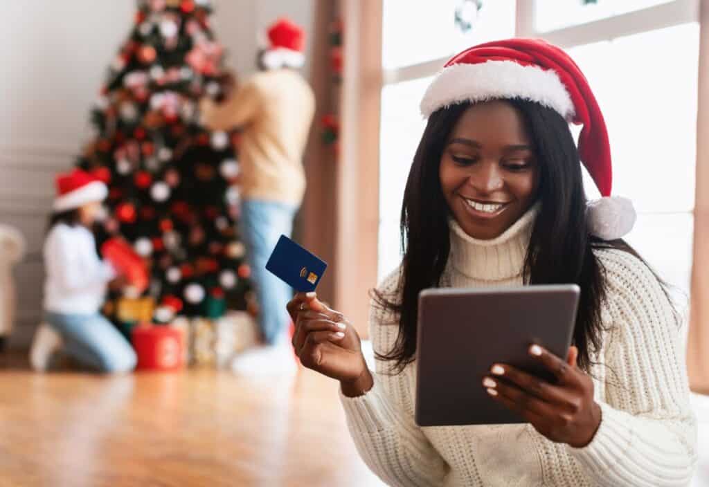 A woman in a Santa hat holds a tablet and credit card, smiling at the fantastic Black Friday deals. In the background, two people decorate a Christmas tree, adding to the festive cheer.