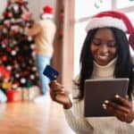 A woman in a Santa hat holds a tablet and credit card, smiling at the fantastic Black Friday deals. In the background, two people decorate a Christmas tree, adding to the festive cheer.