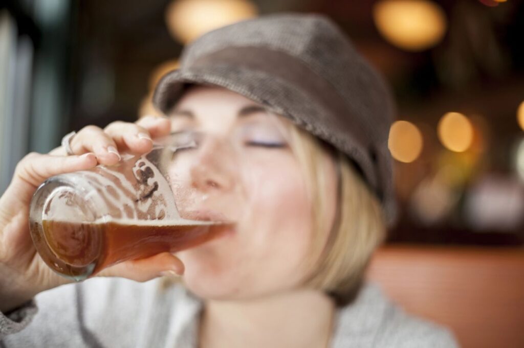 A person wearing a hat drinks from a glass with amber liquid inside, with a blurred background.