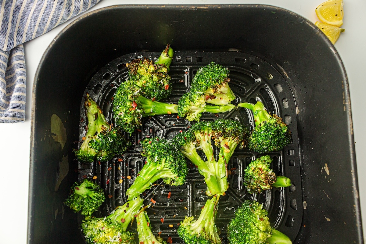 Broccoli florets with seasoning in an air fryer basket, some lemon slices and a striped cloth nearby.