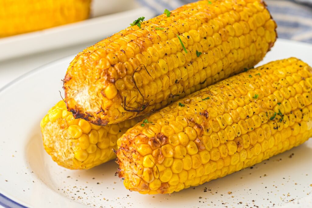 Three grilled corn cobs on a white plate, seasoned with pepper and garnished with herbs.