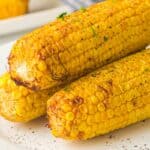 Three grilled corn cobs on a white plate, seasoned with pepper and garnished with herbs.