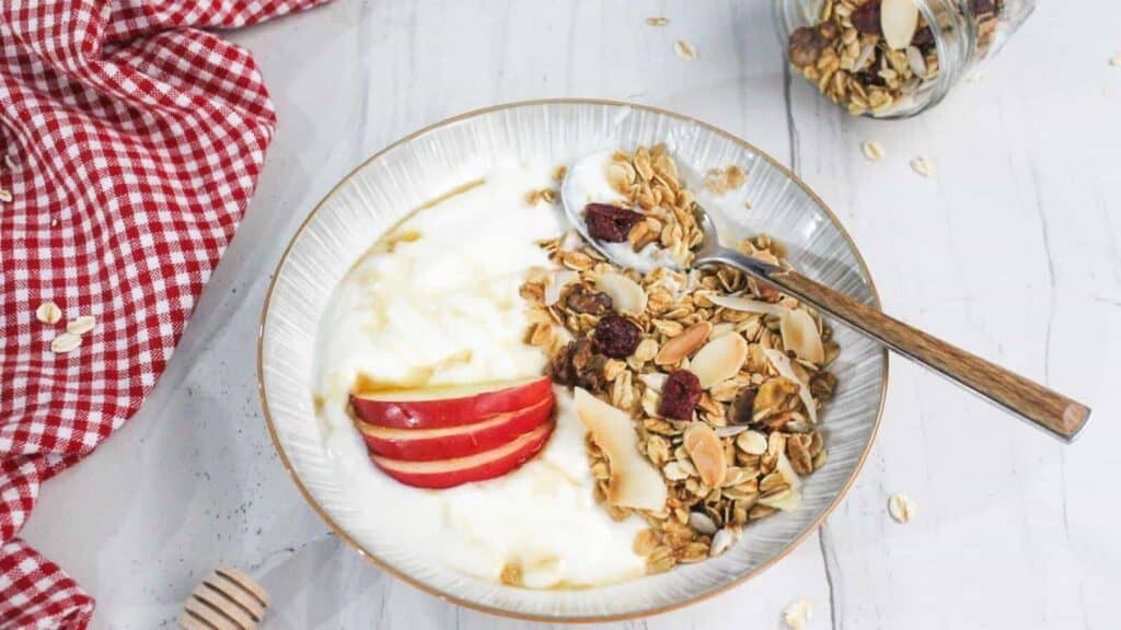 A bowl of yogurt topped with granola, sliced apples, nuts, and honey on a spoon. A red checkered cloth and a jar of granola are in the background.