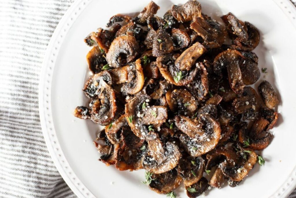 A white plate filled with sautéed mushrooms, garnished with herbs and grated cheese, on a striped cloth background.