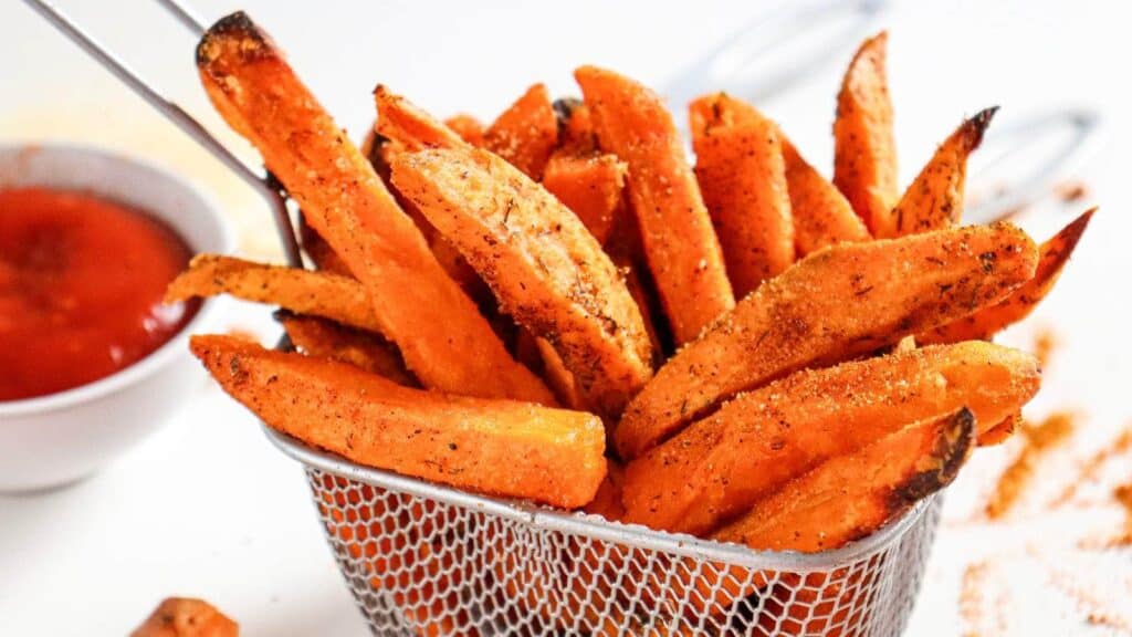 A basket of seasoned sweet potato fries with a small bowl of ketchup in the background.