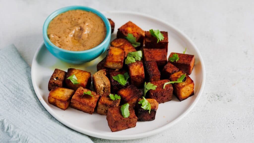 A plate of seasoned, cubed tofu garnished with fresh herbs, accompanied by a bowl of brown dipping sauce.