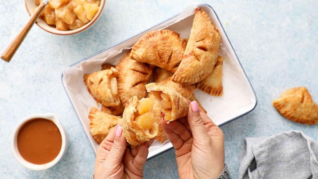 Hands holding a broken apple turnover, revealing the filling. More turnovers are in a tray, with a bowl of apple filling and a cup of caramel sauce nearby. A napkin is on the side.