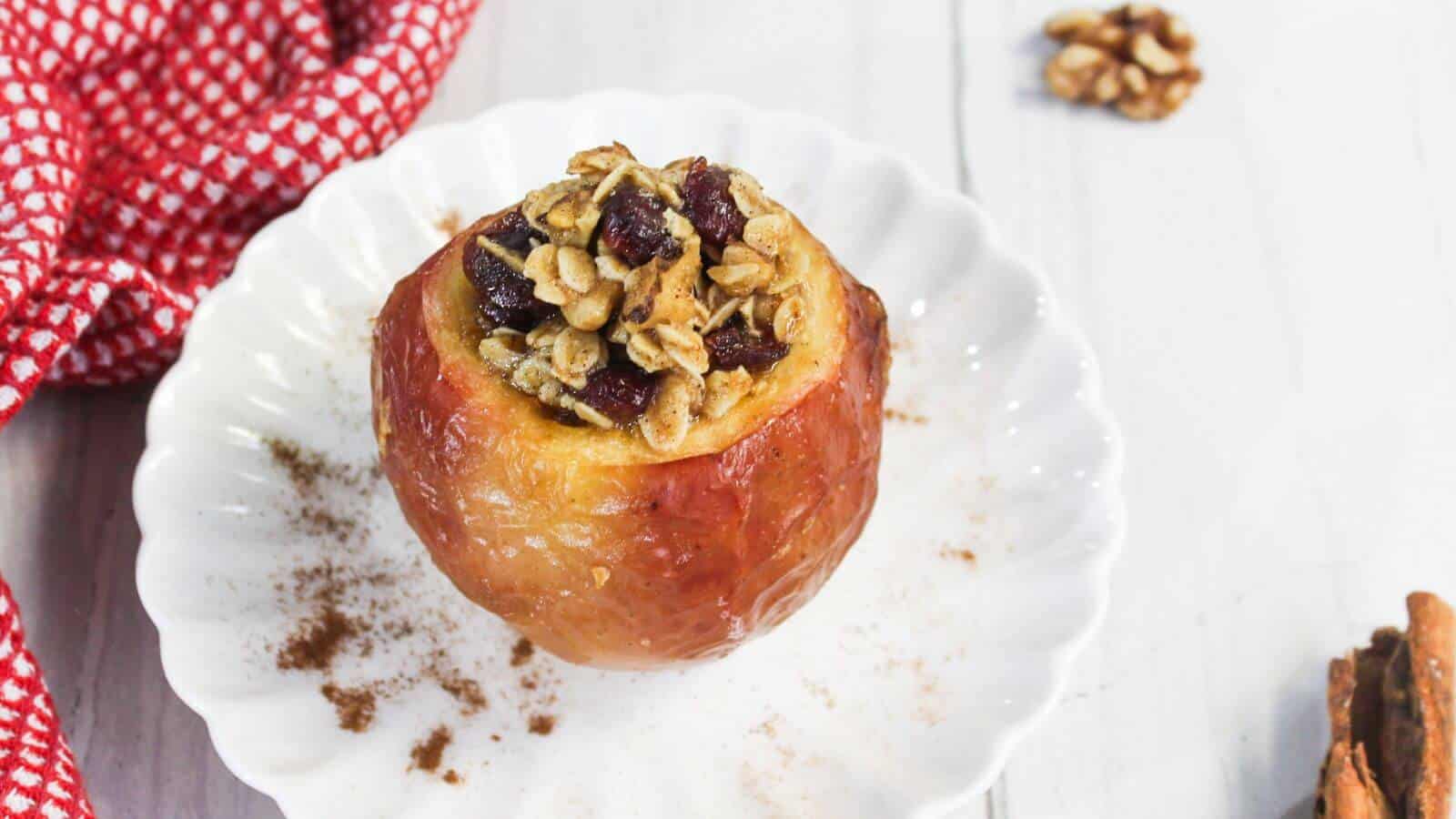 Baked apple on a white plate filled with oats, nuts, and cranberries, with cinnamon sprinkled around. A red checkered cloth is partially visible on the side.