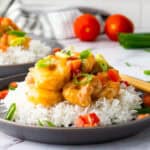 Plate of rice topped with shrimp in sauce, garnished with chopped tomatoes and green onions, with chopsticks on the side. Another similar plate and vegetables in the background.