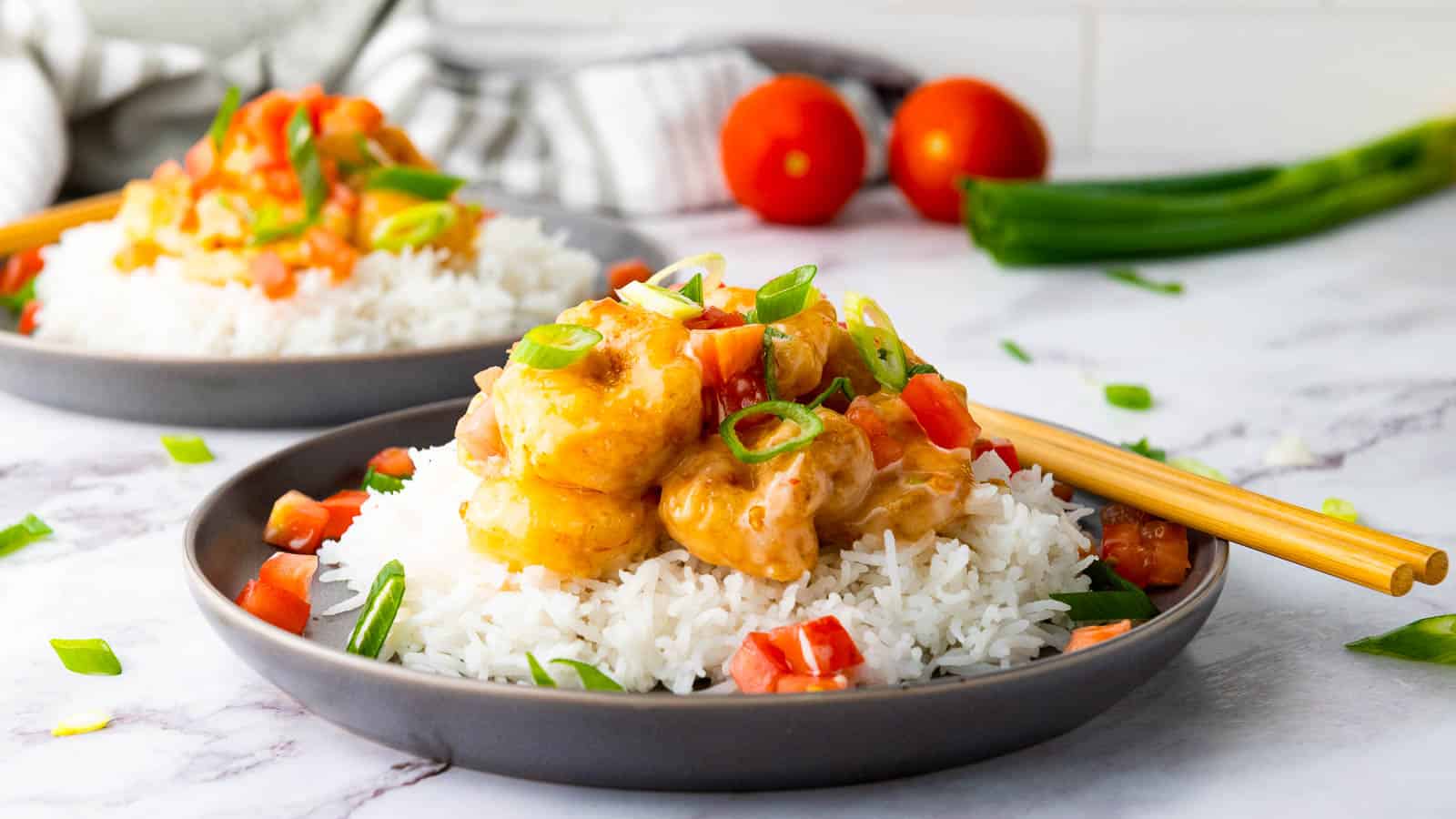 Plate of rice topped with shrimp in sauce, garnished with chopped tomatoes and green onions, with chopsticks on the side. Another similar plate and vegetables in the background.