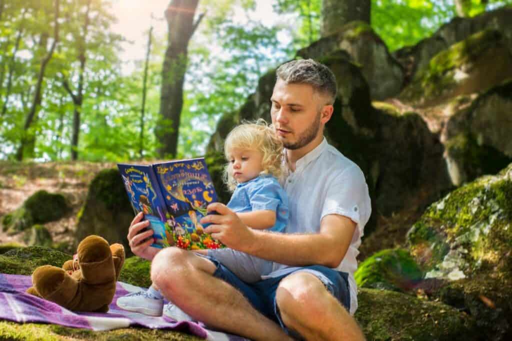 A man and child sit on a blanket outdoors, reading a colorful book together. A teddy bear sits nearby. The setting is a wooded area with sunlight filtering through the trees.