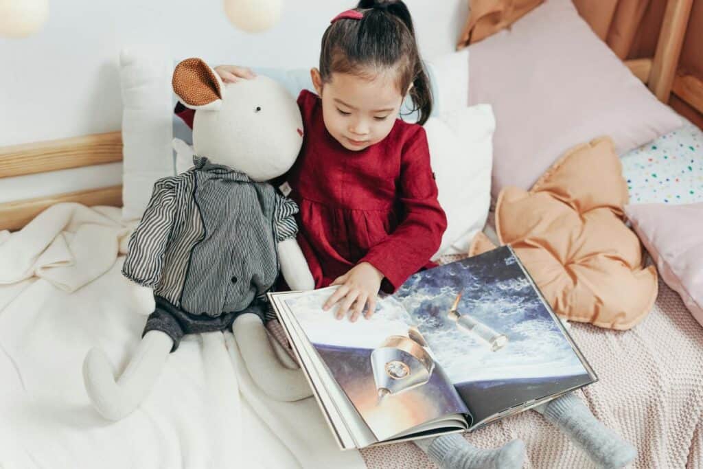 A young girl in a red dress reads a picture book about space, sitting on a bed with a stuffed toy rabbit by her side.