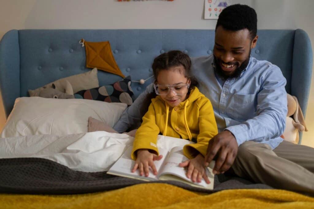 A child wearing glasses reads a book with an adult on a bed. The adult is smiling and pointing at the book. The bed has a blue headboard and various bedding.