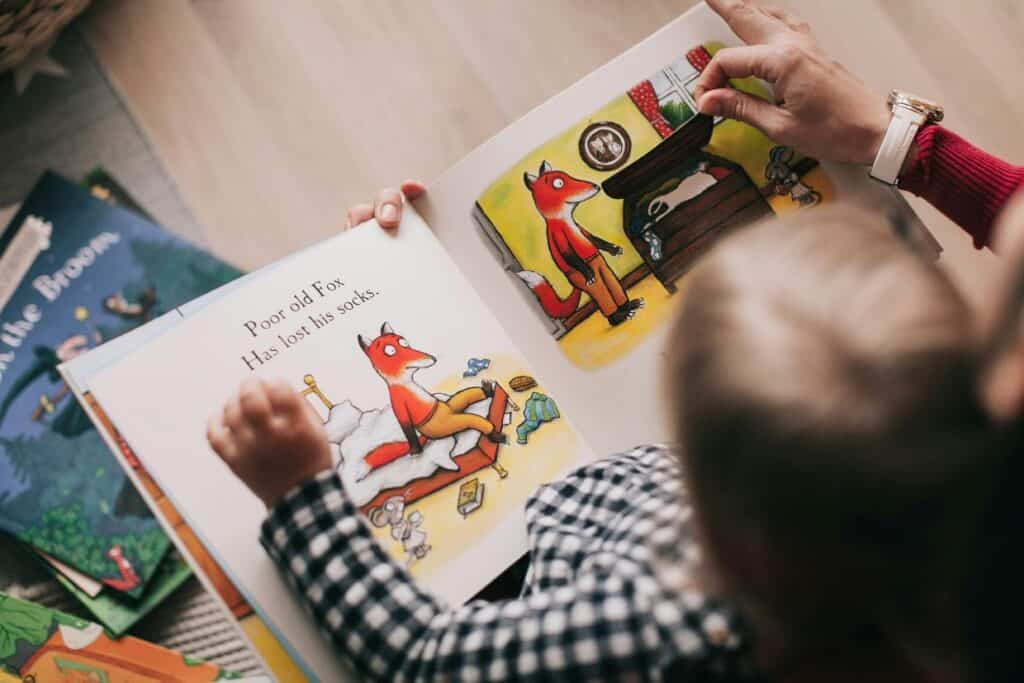 A child reads a picture book about a fox with someone. The book shows illustrations of a fox searching for lost socks. Other books are nearby on the floor.