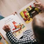 A child reads a picture book about a fox with someone. The book shows illustrations of a fox searching for lost socks. Other books are nearby on the floor.