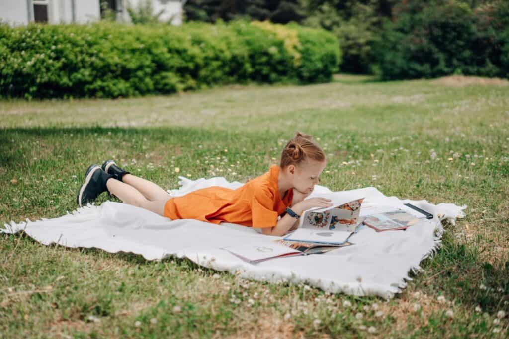 Girl in an orange dress lies on a white blanket in a grassy area, reading a book and looking intently.