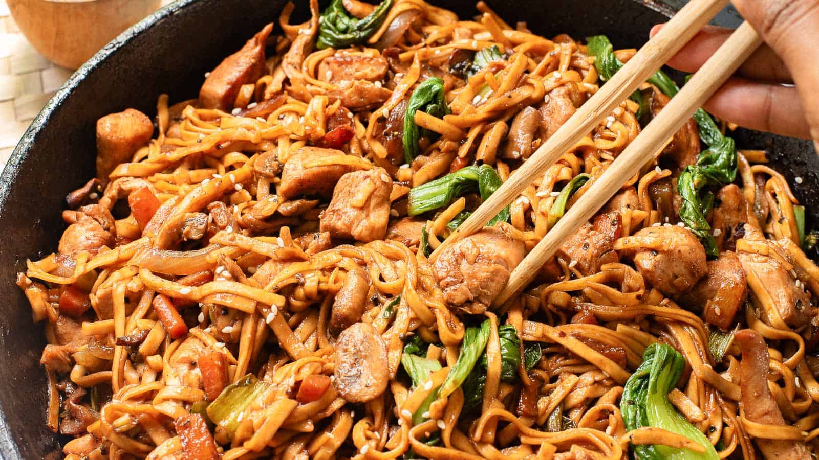 A skillet filled with stir-fried noodles, chicken pieces, and vegetables being picked up with chopsticks.