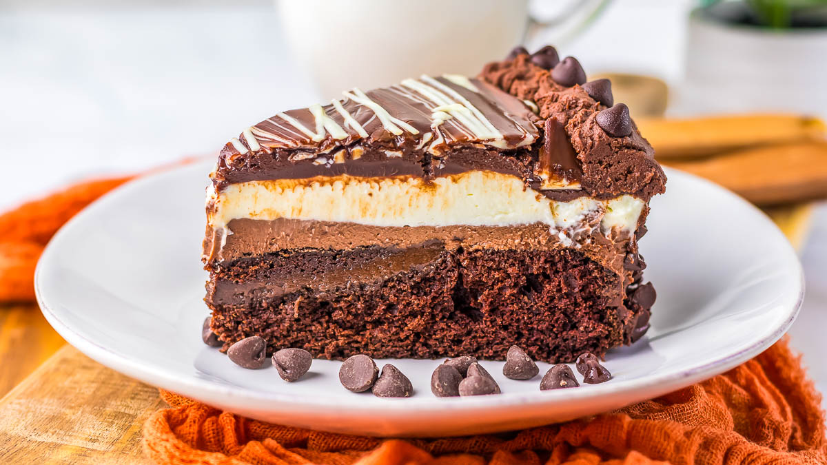 A slice of multi-layered chocolate cake with frosting, chocolate chips, and decorative drizzle on a white plate.