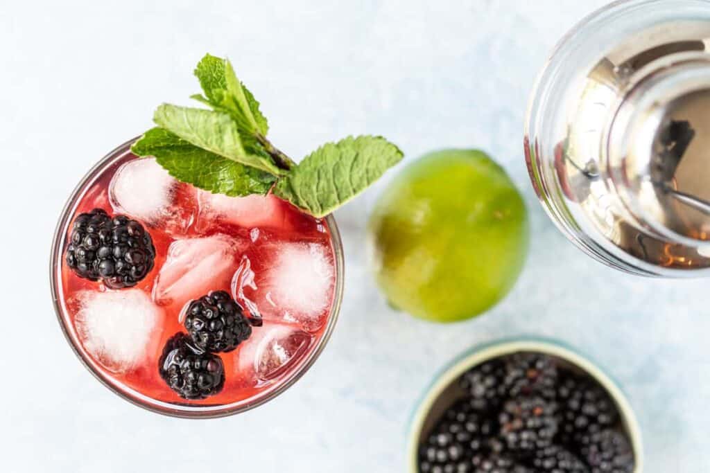 A glass of iced blackberry cocktail garnished with mint, next to a whole lime and a bowl of blackberries.